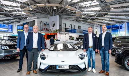 Vier Männer in Anzügen stehen in einem modernen Autohaus neben einem weißen Ford GT Sportwagen im Mittelpunkt des Showrooms.