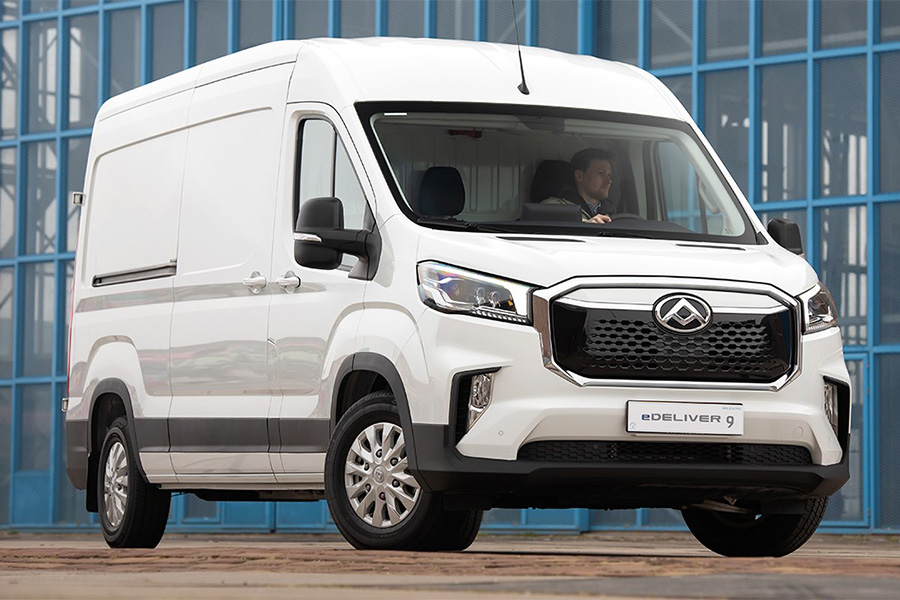 Ein weißer Maxus eDeliver 9 Transporter steht vor einem modernen Gebäude mit blauen Glasfassaden. Der Transporter hat ein markantes Design mit großem Kühlergrill und LED-Scheinwerfern.