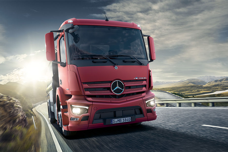Ein roter Mercedes-Benz Actros Lkw fährt bei Sonnenaufgang auf einer kurvigen Bergstraße entlang, umgeben von einer weiten, hügeligen Landschaft.