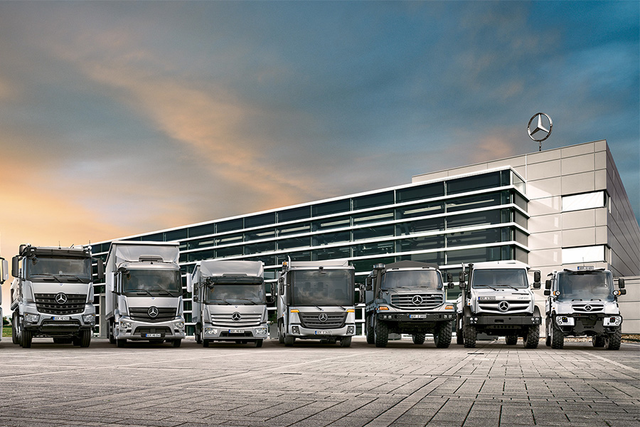 Eine Reihe von acht Mercedes-Benz Nutzfahrzeugen, darunter Lkw und Unimog-Modelle, steht vor einem modernen Bürogebäude mit Mercedes-Stern auf dem Dach bei Sonnenuntergang.