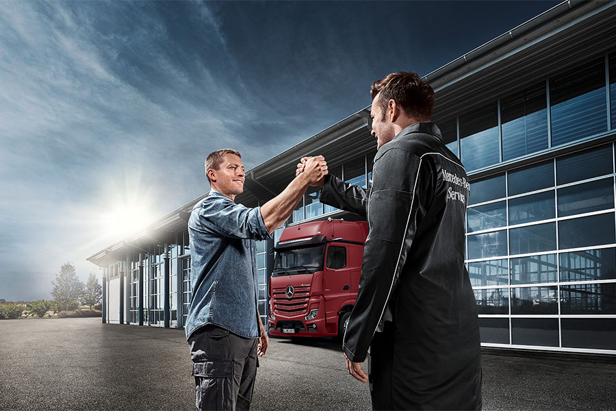 Zwei Männer – ein Lkw-Fahrer und ein Mercedes-Benz Servicemitarbeiter – geben sich vor einer modernen Werkstatt mit Glasfassade und einem roten Mercedes-Benz Lkw im Hintergrund einen respektvollen Handschlag.
