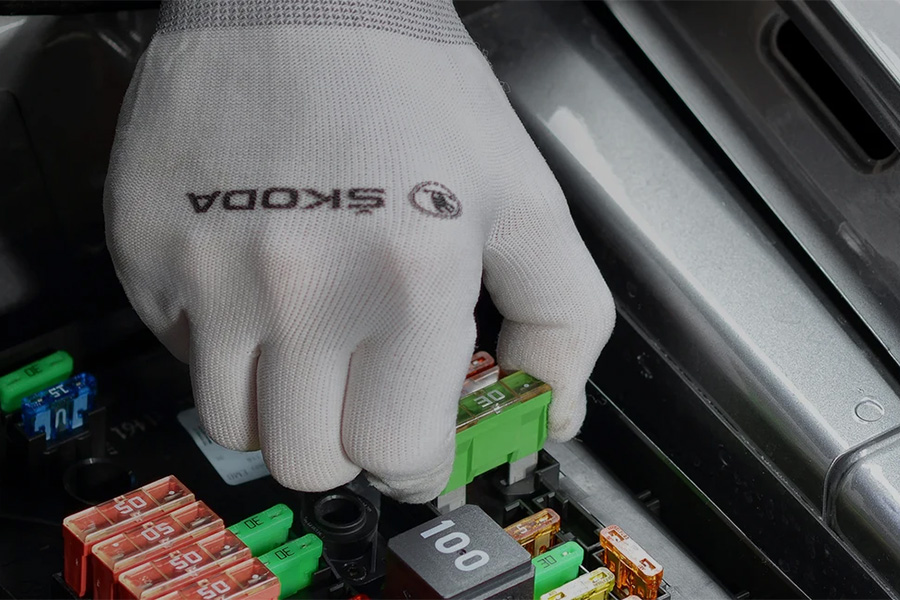 Eine Hand mit einem weißen Handschuh, auf dem das ŠKODA-Logo zu sehen ist, greift nach einer grünen Sicherung in einem Auto-Sicherungskasten. Umgeben sind die Sicherungen von verschiedenen anderen Sicherungen in Rot, Blau, Orange und Schwarz.