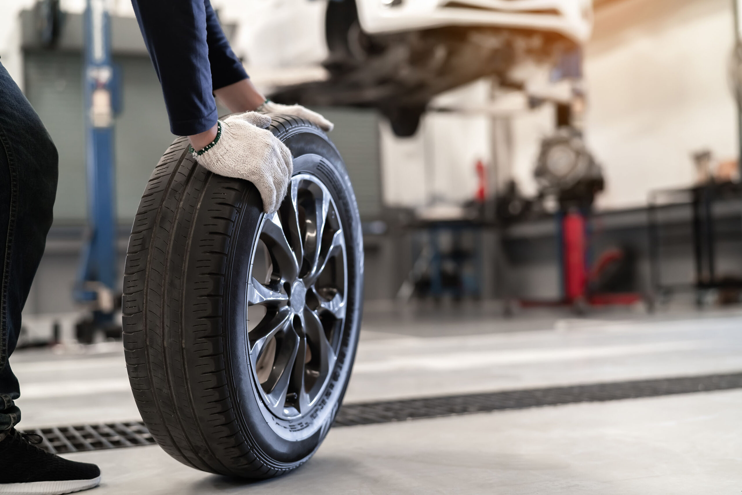Ein Mechaniker mit Handschuhen hält einen Autoreifen aufrecht in einer Werkstatt. Im Hintergrund ist ein Auto auf einer Hebebühne zu sehen, das zur Wartung angehoben wurde.