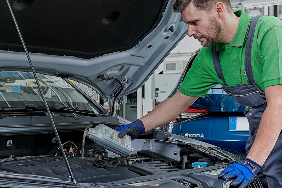 Ein Škoda-Servicetechniker in grüner Arbeitskleidung füllt Motoröl in ein Fahrzeug ein. Die Motorhaube ist geöffnet, im Hintergrund ist ein weiterer blauer Škoda sichtbar – die Szene zeigt eine professionelle Wartungssituation in einer Werkstatt.