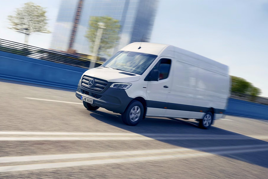 Ein weißer Mercedes-Benz eSprinter Kastenwagen fährt dynamisch über eine städtische Brücke, im Hintergrund moderne Hochhäuser und verschwommene Bewegungslinien, die Geschwindigkeit andeuten.