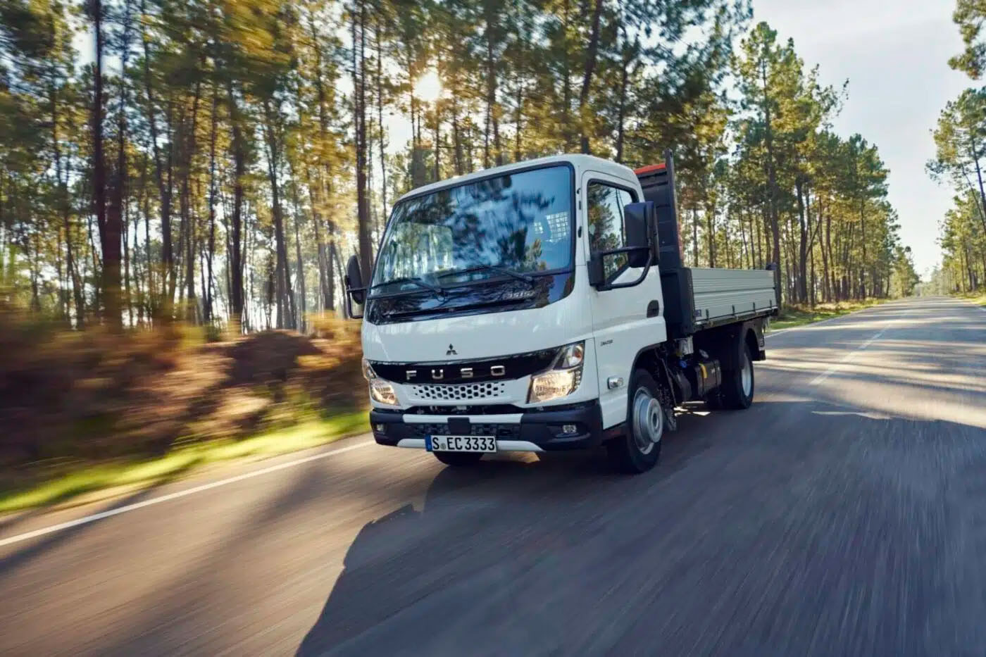 Ein weißer FUSO Canter LKW fährt dynamisch auf einer Landstraße durch einen sonnenbeschienenen Wald.