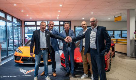 Fünf Männer in Business-Outfits stehen in einem Autohaus vor zwei Sportwagen (gelb und rot) und legen ihre Hände gemeinsam in die Mitte – Symbol für Zusammenarbeit oder Vertragsabschluss.