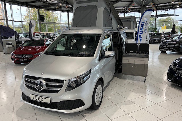 Silberner Mercedes-Benz Marco Polo mit ausgefahrenem Aufstelldach und geöffneter Seitenküche steht in einem Autohaus zwischen weiteren Fahrzeugen.