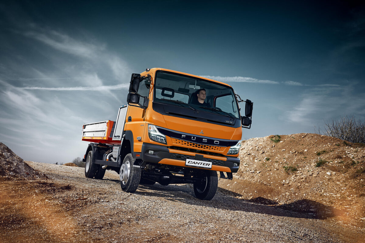 Ein orangefarbener FUSO Canter LKW fährt über einen unebenen Schotterweg in einer Offroad-Umgebung unter blauem Himmel.