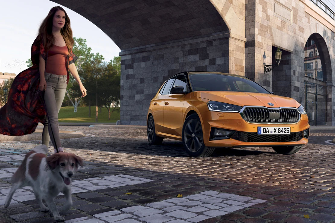 Eine Frau mit Hund geht an einem orangefarbenen Škoda-Kompaktwagen vorbei, der unter einem steinernen Brückenbogen auf Kopfsteinpflaster steht.