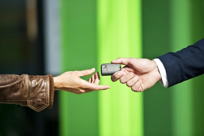Eine Person in brauner Lederjacke nimmt von einer Person im Anzug einen Autoschlüssel entgegen, vor einem Hintergrund mit grünen vertikalen Streifen.