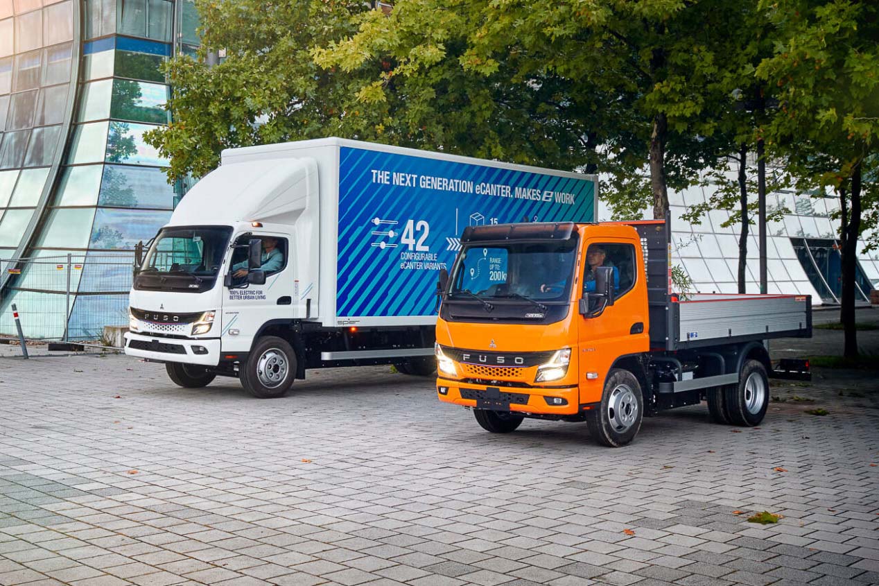 Zwei FUSO Canter LKWs nebeneinander auf einem gepflasterten Platz – ein weißer eCanter mit großem Kastenaufbau und ein orangefarbener Pritschenwagen, im Hintergrund moderne Glasarchitektur und Bäume.