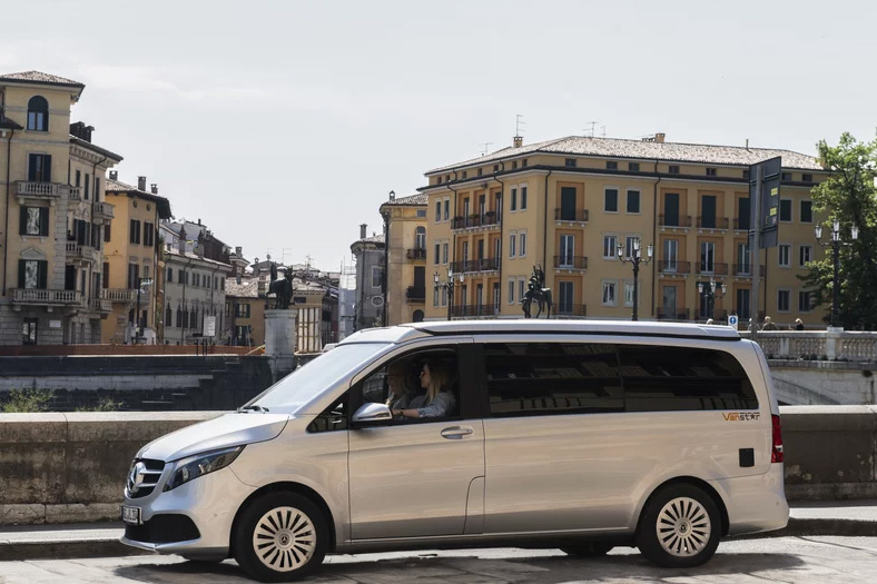 Silberner Mercedes-Benz Vanster Campervan steht am Ufer einer Stadt mit historischen Gebäuden im Hintergrund, eine Frau sitzt am Steuer.
