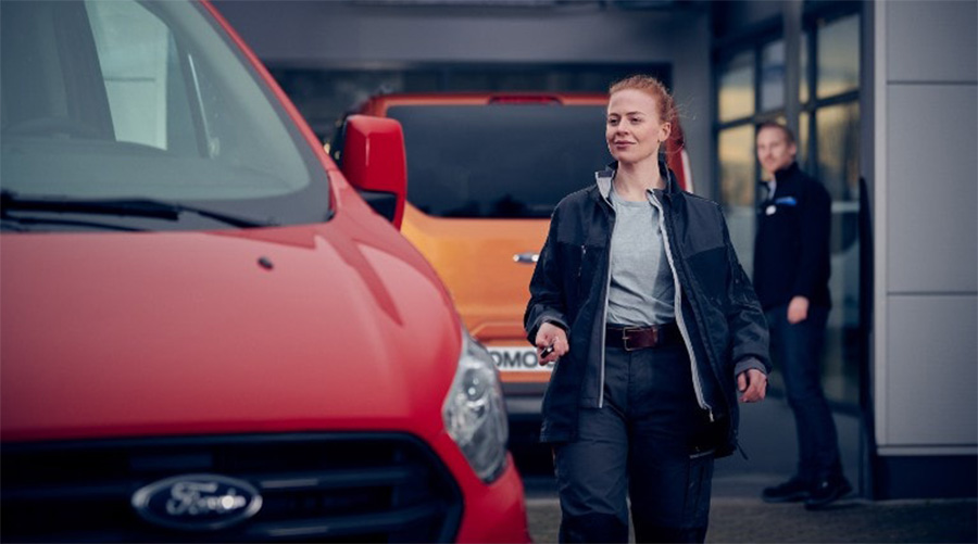 Junge Frau in Arbeitskleidung geht lächelnd an einem roten Ford Nutzfahrzeug vorbei, im Hintergrund ein orangefarbener Transporter und ein Mann im Anzug vor einem Autohaus.