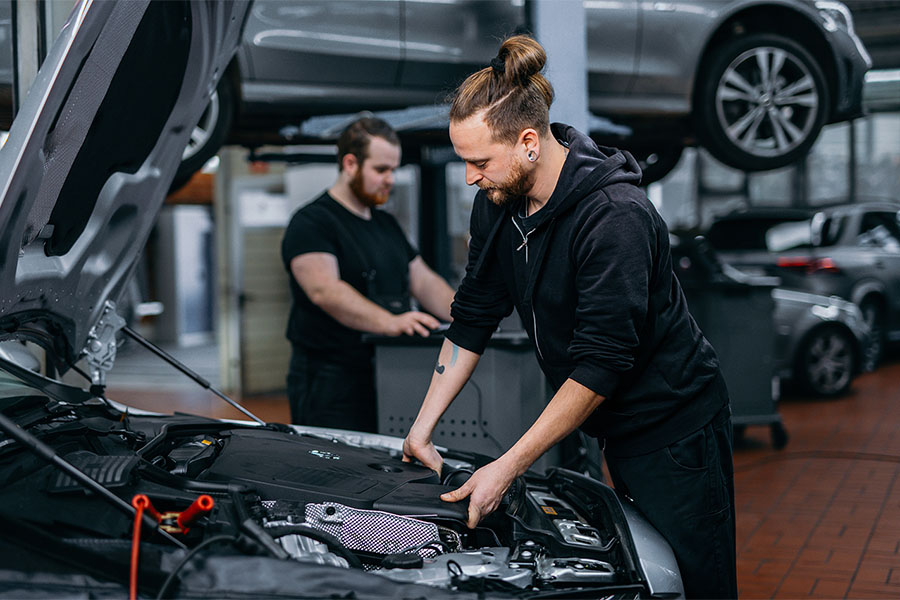 Das Bild zeigt eine Werkstattszene mit zwei Kfz-Mechatronikern. Der Fokus liegt auf dem vorderen Techniker, der an einem Motorraum arbeitet – vermutlich bei einer Diagnose, Wartung oder Reparatur. Er trägt legere, dunkle Arbeitskleidung und wirkt konzentriert.