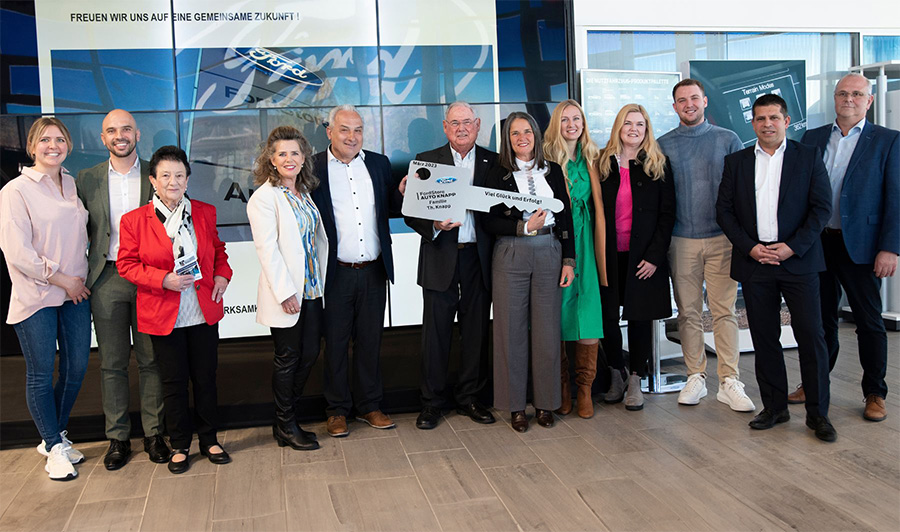 Gruppenfoto von 14 Personen bei einer symbolischen Schlüsselübergabe in einem Ford-Autohaus – festlich gekleidet, fröhliche Stimmung, großes Ford-Logo im Hintergrund.