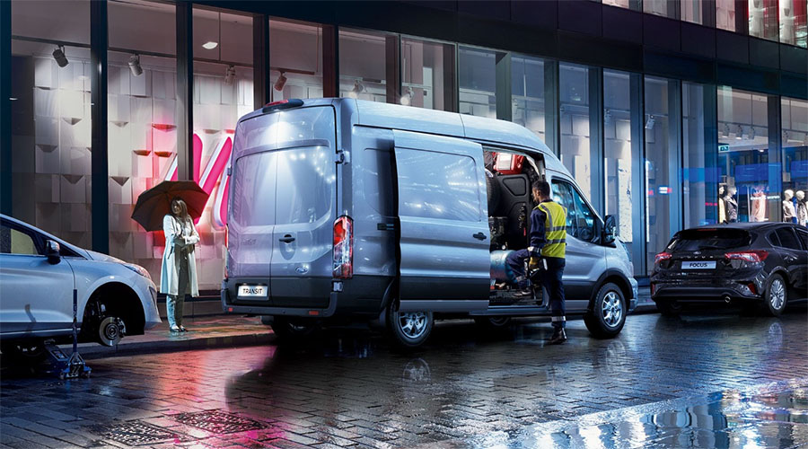 Ford Transit bei nächtlichem Einsatz in der Stadt – Handwerker lädt Werkzeug aus, Passantin mit Regenschirm geht vorbei.