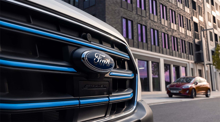 Nahaufnahme des Ford-Logos am Kühlergrill eines E-Transit vor moderner Architektur, im Hintergrund ein geparkter roter Ford Fiesta.
