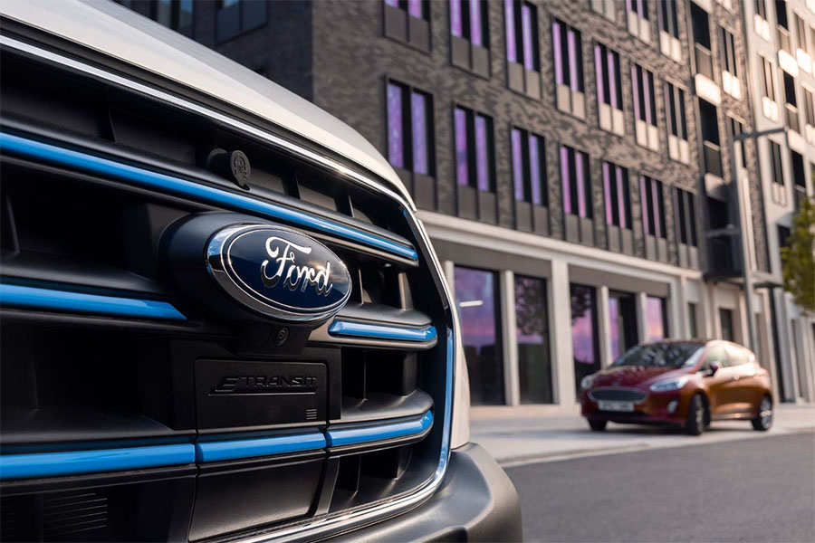 Nahaufnahme des Ford-Logos am Kühlergrill eines E-Transit vor moderner Architektur, im Hintergrund ein geparkter roter Ford Fiesta.