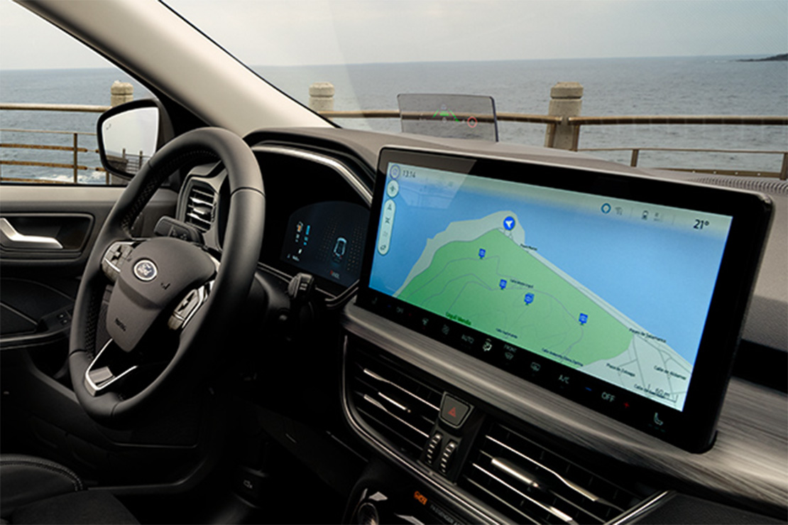 Innenraum eines Ford-Fahrzeugs mit digitalem Cockpit und großem Navigationsdisplay, geparkt an einer Küstenstraße mit Blick auf das Meer.