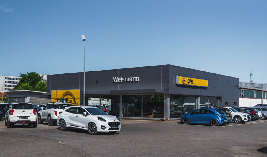 Autohaus Weisßmann mit Opel-Logo und moderner Glasfassade. Vor dem Gebäude stehen mehrere Fahrzeuge auf dem Parkplatz, darunter ein weißer Ford Puma im Vordergrund.
