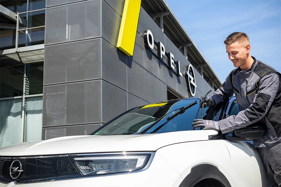 Ein Opel-Servicetechniker wechselt freundlich die Scheibenwischer eines weißen Opel SUV vor einem modernen Autohaus mit Opel-Logo – zuverlässiger Kundenservice und Fahrzeugpflege.