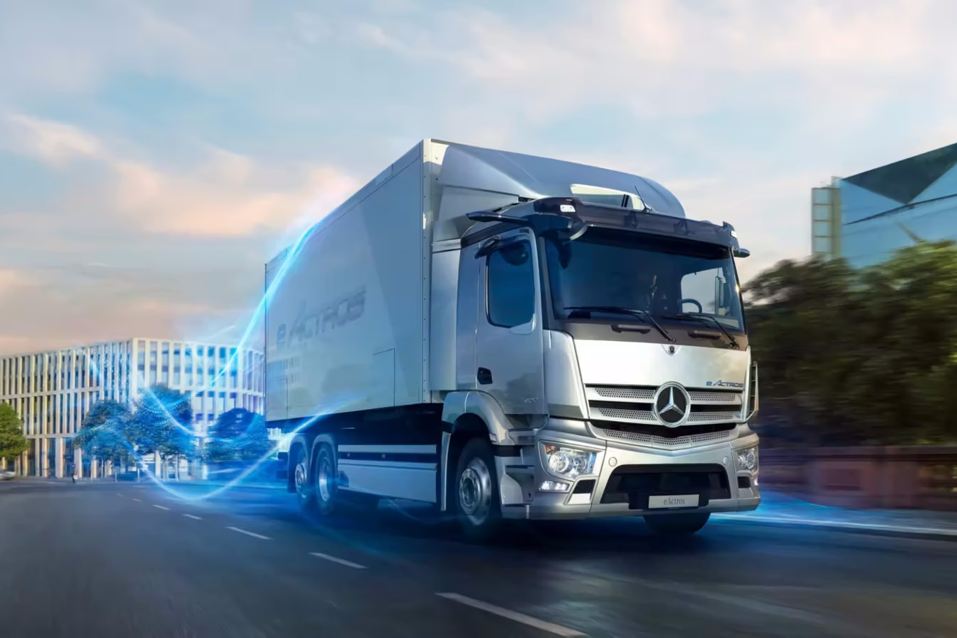 Moderner Mercedes-Benz eActros Elektro-LKW fährt dynamisch durch eine urbane Straße bei Sonnenuntergang. Blaue Energieeffektlinie symbolisiert elektrische Antriebskraft. Im Hintergrund moderne Bürogebäude und Bäume.