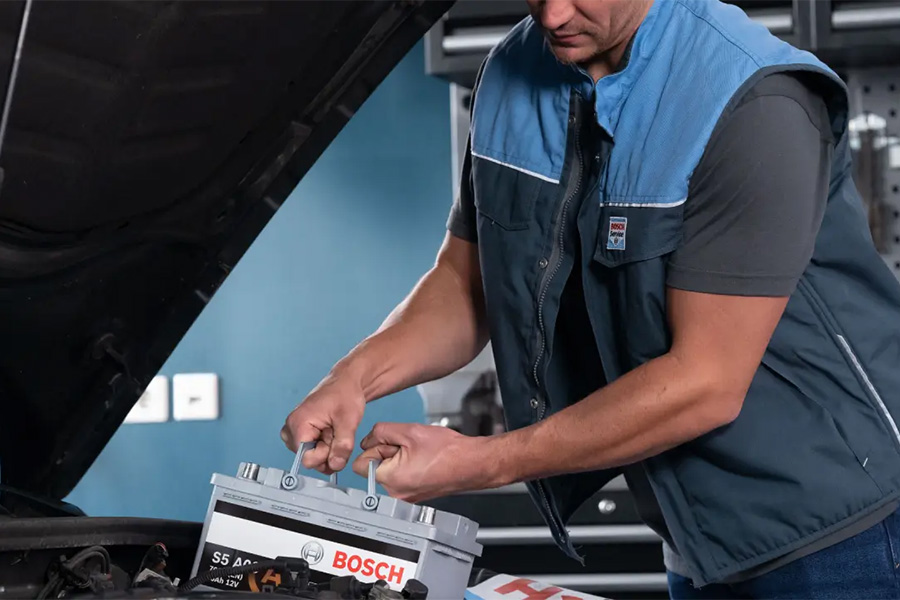 Kfz-Mitarbeiter in Werkstattkleidung setzt eine Bosch-Autobatterie in den Motorraum eines Fahrzeugs ein. Die Motorhaube ist geöffnet.