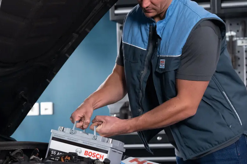 Kfz-Mitarbeiter in Werkstattkleidung setzt eine Bosch-Autobatterie in den Motorraum eines Fahrzeugs ein. Die Motorhaube ist geöffnet.