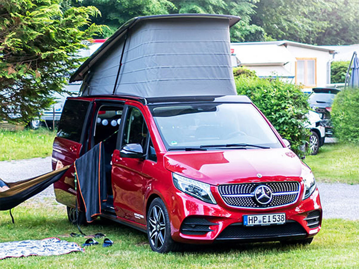 Roter Mercedes-Benz Campingbus mit aufgeklapptem Aufstelldach steht auf einem Campingplatz. Die seitliche Schiebetür ist geöffnet, eine Hängematte hängt neben dem Fahrzeug.