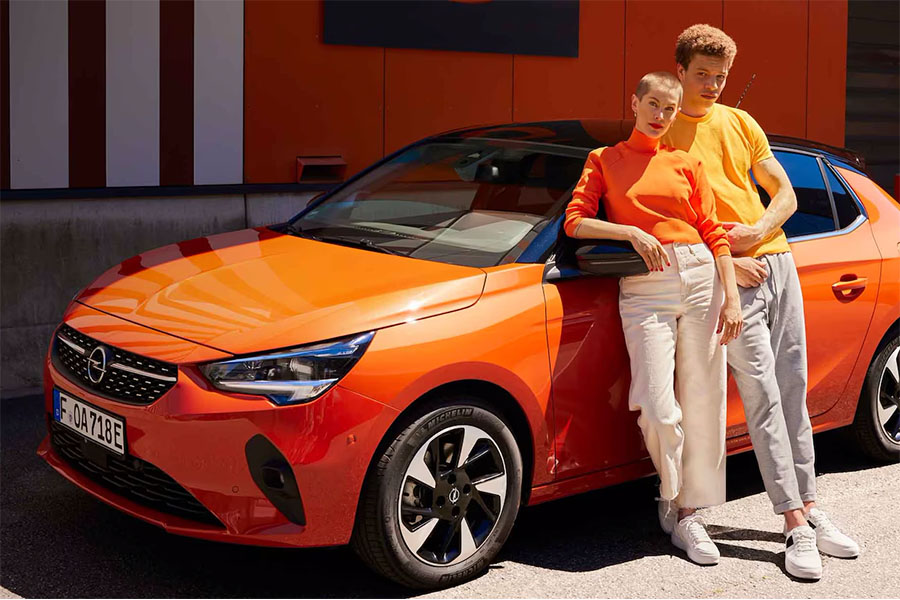 Zwei junge Personen lehnen an einem orangefarbenen Opel Corsa vor einer farblich abgestimmten Gebäudewand. Beide tragen sportlich-leuchtende Kleidung in Orange- und Gelbtönen.