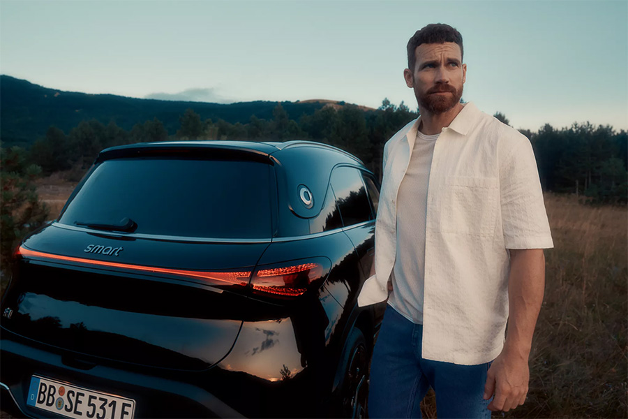 Mann in heller Kurzarmjacke steht neben einem schwarzen Smart SUV in einer offenen Waldlandschaft bei Sonnenuntergang. Das Fahrzeug ist von hinten zu sehen, Rückleuchten leuchten leicht.