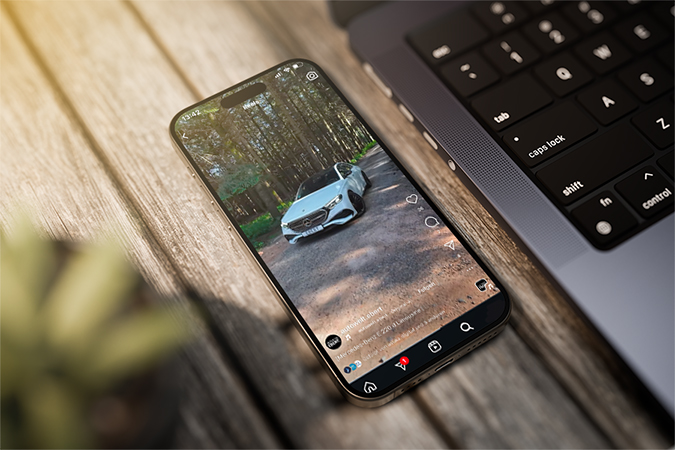Smartphone liegt auf einem Holztisch neben einer Laptop-Tastatur. Auf dem Display ist ein Instagram-Video eines silbernen Mercedes-Benz zu sehen, der auf einem Waldweg steht.