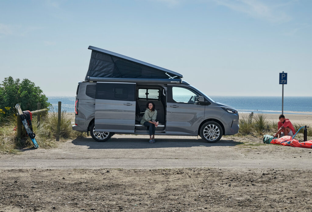 Ein grauer Ford Transit Nugget seitlich zu sehen mit Aufstelldach vor einer Meeresküste.
