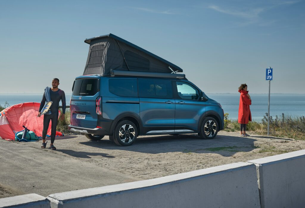 Ein blauer Ford Transit Nugget Custom seitliche zu sehen, links und rechts daovn steht jeweils eine Person, im Hintergrund sieht man eine Küste.