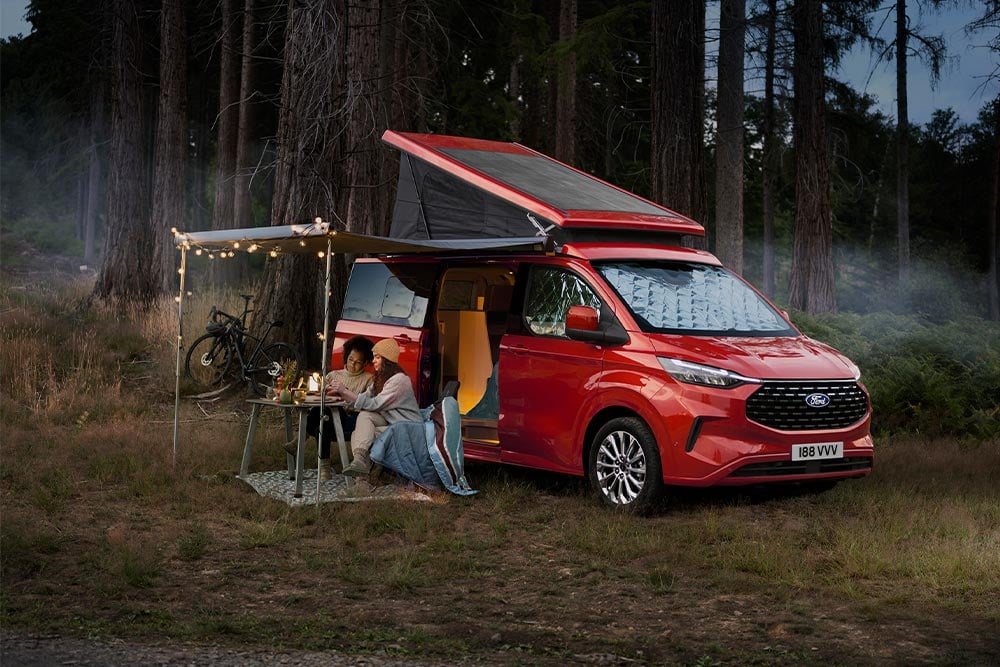 Ein roter Ford Transit Custom Nugget mit Aufstelldach und ausgelassener Markise, darunter sitzen zwei Personen an einem Campingtisch mitten im Wald.