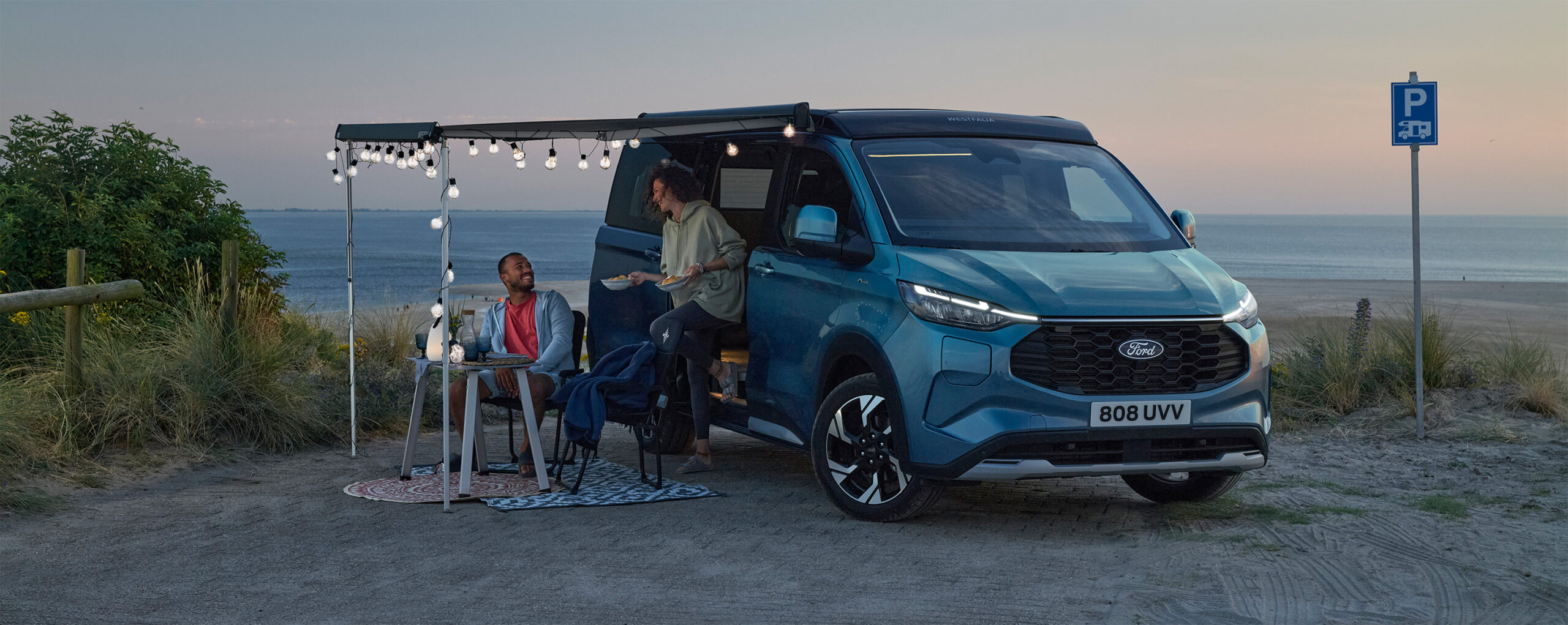 Ein blauer Ford Transit Nugget Custom seitliche zu sehen, links und rechts daovn steht jeweils eine Person, im Hintergrund sieht man eine Küste.