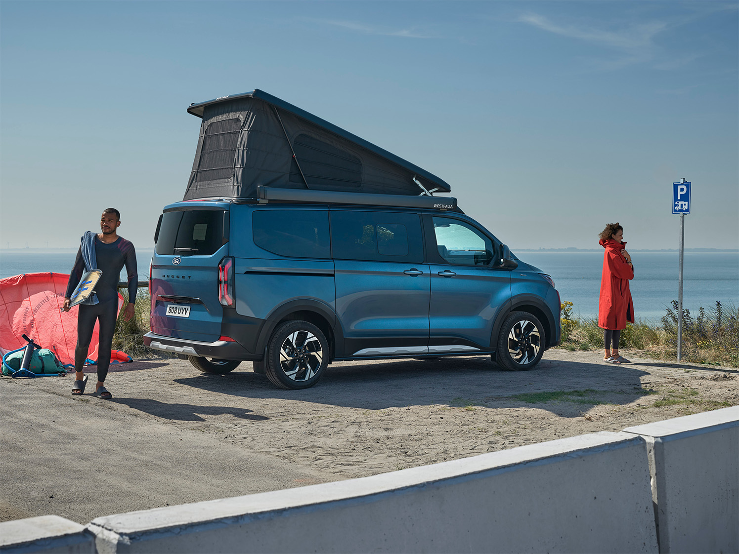 Ein blauer Ford Transit Nugget Custom seitliche zu sehen, links und rechts daovn steht jeweils eine Person, im Hintergrund sieht man eine Küste.