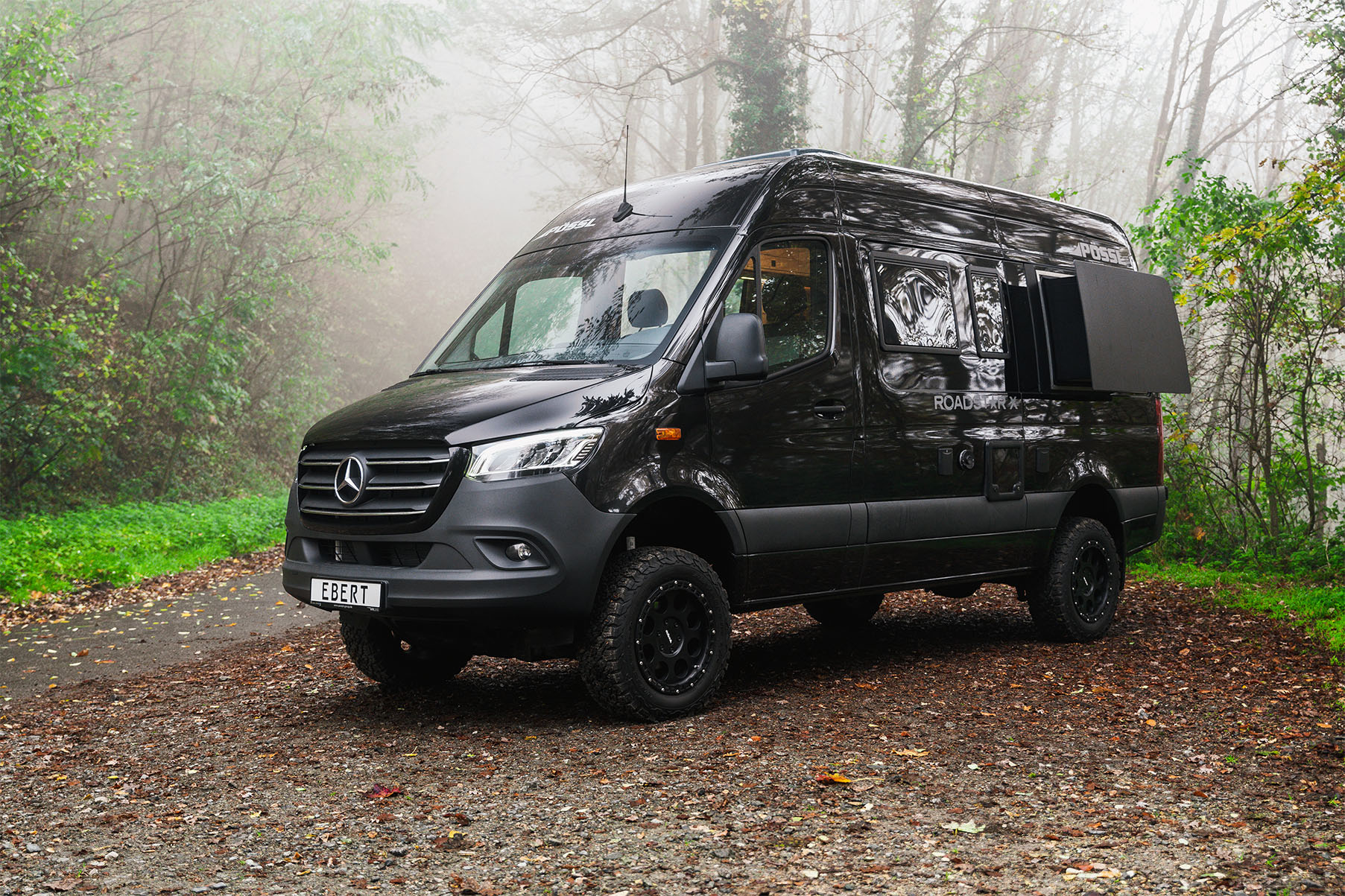 Schwarzer Mercedes-Benz Kastenwagen der Marke Pössl mit Offroad-Bereifung und geöffnetem Seitenfenster, geparkt auf einem Waldweg bei Nebel; Kennzeichen trägt den Schriftzug „EBERT“.