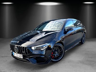 Front- und Seitenansicht eines schwarzen Mercedes-AMG CLA 45 S Shooting Brake mit Panamericana-Kühlergrill, LED-Scheinwerfern, roten Bremssätteln und schwarzen AMG-Felgen. Das Fahrzeug steht in einem Studio mit Spotbeleuchtung auf grauem Betonboden.
