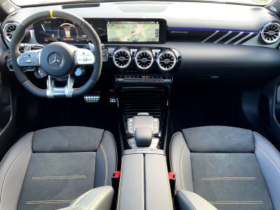 Cockpit-Ansicht eines Mercedes-AMG mit digitalem Kombiinstrument, Navigationsbildschirm, beleuchteter Zierleiste und drei runden Lüftungsdüsen in der Mittelkonsole. AMG Performance-Lenkrad mit gelbem Mittenmarker und Sportsitze mit gelben Ziernähten sichtbar.